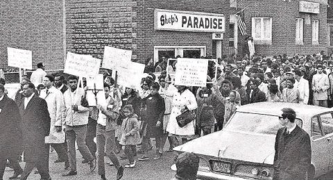 3,000 people marched on Clarissa Street following the funeral of Martin Luther King Jr. Shep's Paradise, another club, is pictured in the background. The demonstrators are holding signs, most are African Americans. A 1950's car is in the foreground.