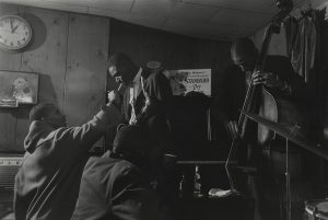 One man leans over to have a cigarette lit. A man on his right is playing an upright bass,