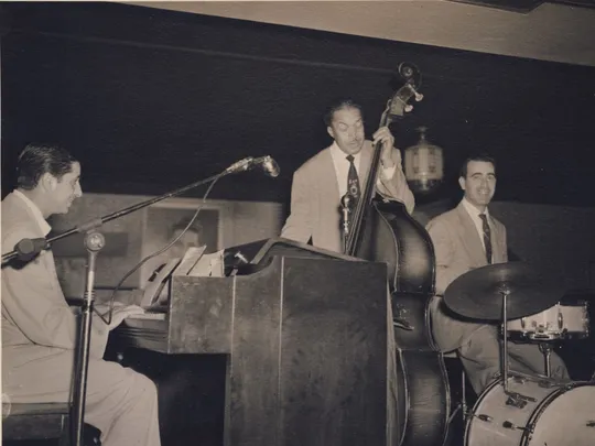 A black and white photo depicts a pianist, a bassist, and a drummer preparing to play.